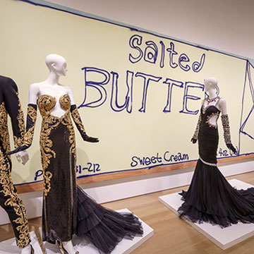 mannequins wearing black and gold designs stand in front of a large painting of salted butter sticks